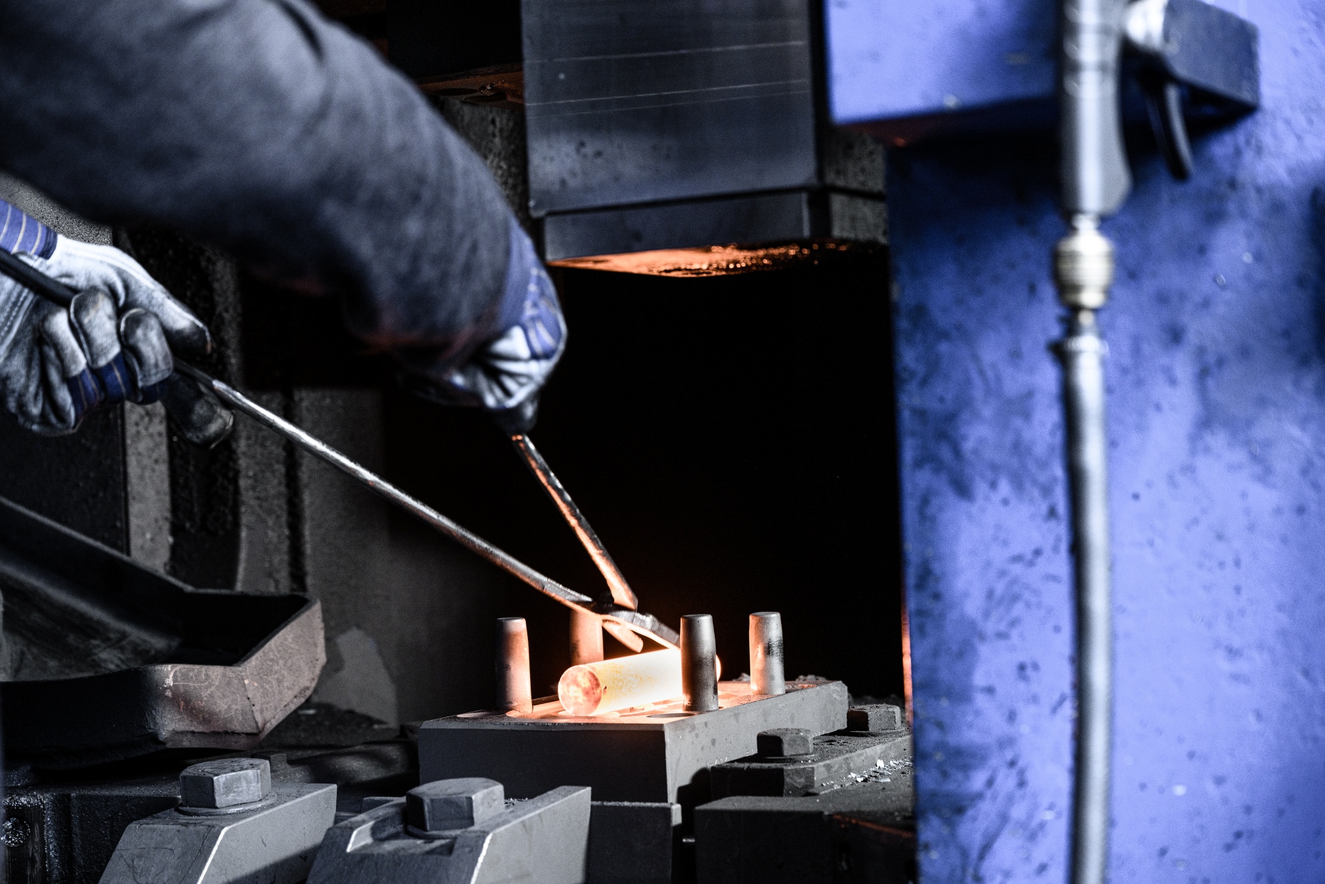 Nahaufnahme eines Produktionsprozesses: Eine geschützte Hand positioniert glühendes Stahl in einer Formpresse in einer Industriehalle.
