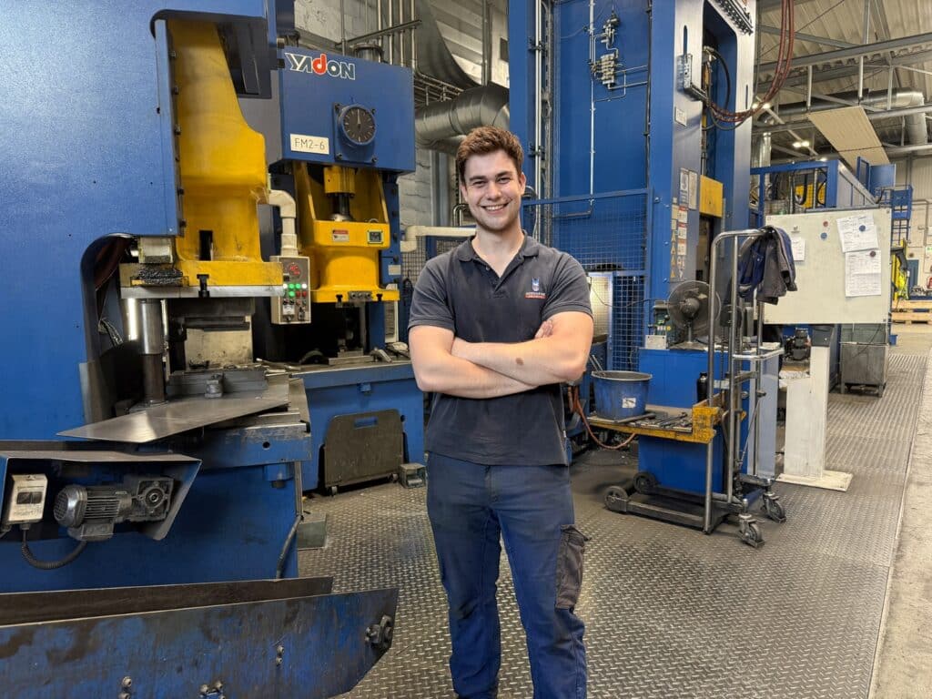 Mitarbeitende Person in Arbeitskleidung steht in einer Produktionshalle vor einer blauen und gelben Industrie‑Presse, umgeben von Maschinen und Fertigungsanlagen.
